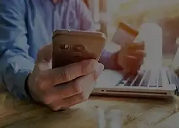 Person holding a smartphone and a payment card while using a laptop for an online transaction.